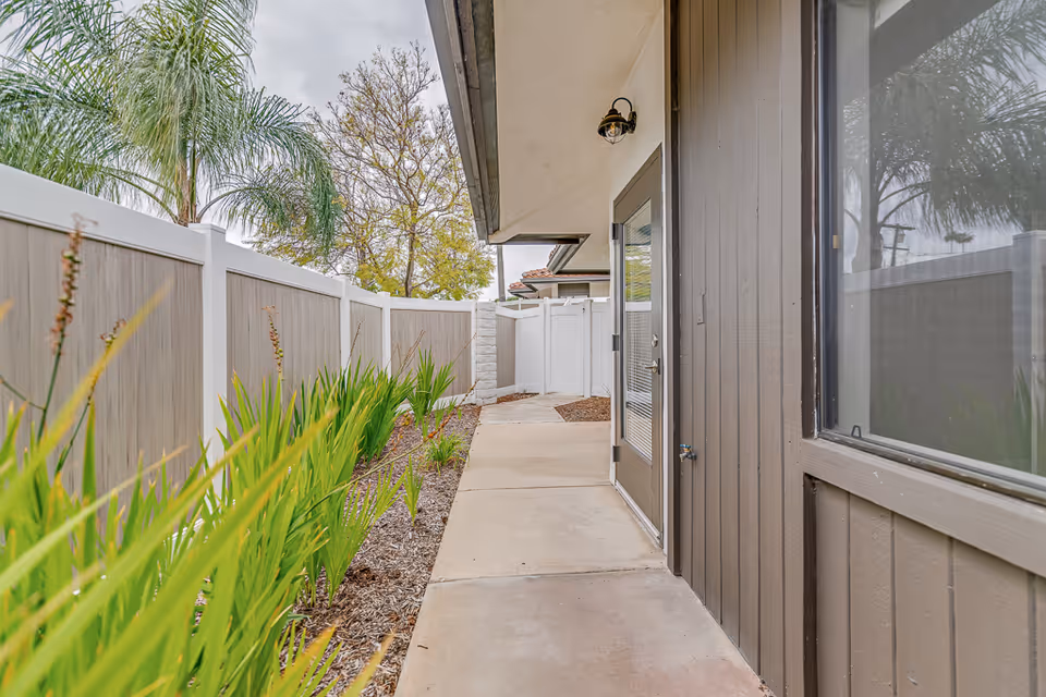 Narrow outdoor walkway beside a brown building with a glass door, white fence, and planted landscaping.