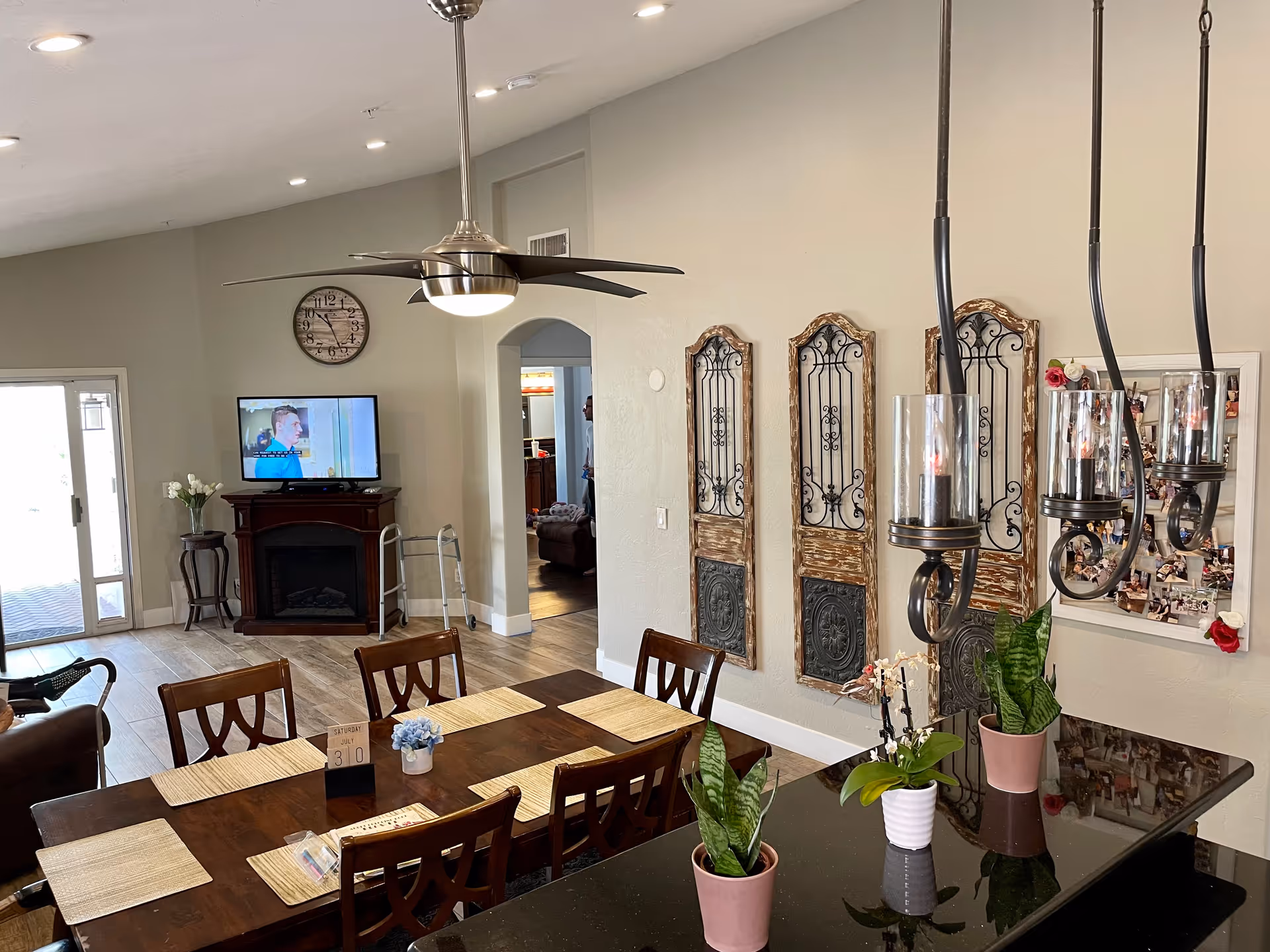 Interior view of a senior living facility common area with a dining table set with placemats and a small flower pot. There are wooden chairs around the table. In the background, a TV is mounted above a fireplace with a walker nearby. The walls are decorated with ornate wooden panels and a bulletin board with photos. Ceiling lights and a modern ceiling fan are visible, along with potted plants on a black countertop in the foreground.