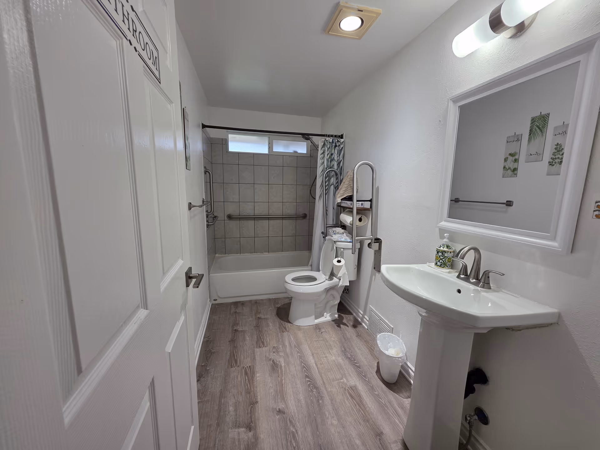 A clean bathroom with a white door labeled 'BATHROOM' on the left. Inside, there is a white toilet with grab bars and a shelf holding towels and toilet paper. A bathtub with tiled walls and a shower curtain is at the back. On the right, there is a white pedestal sink with a soap dispenser and a large mirror above it. The floor has wood-like vinyl flooring and the walls are painted white.