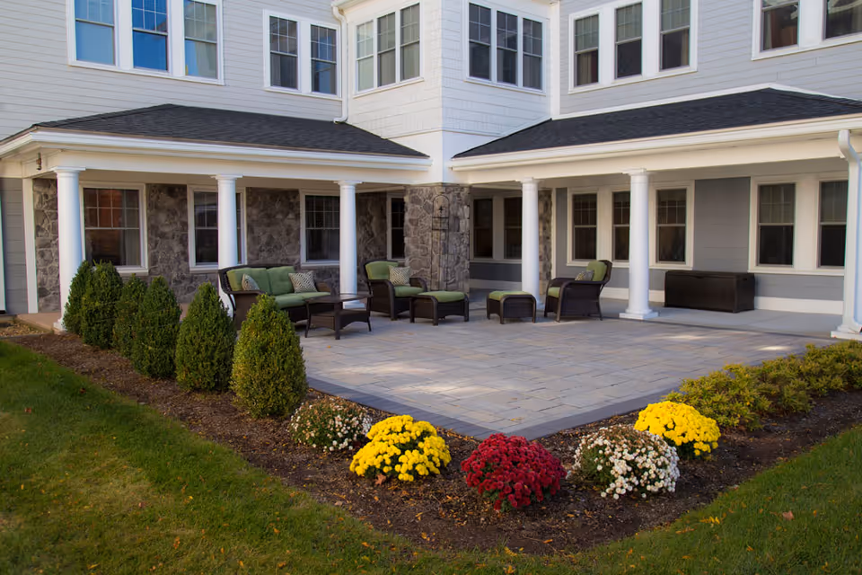 A paved patio with outdoor seating and flower beds in front of a multi-story senior living building.