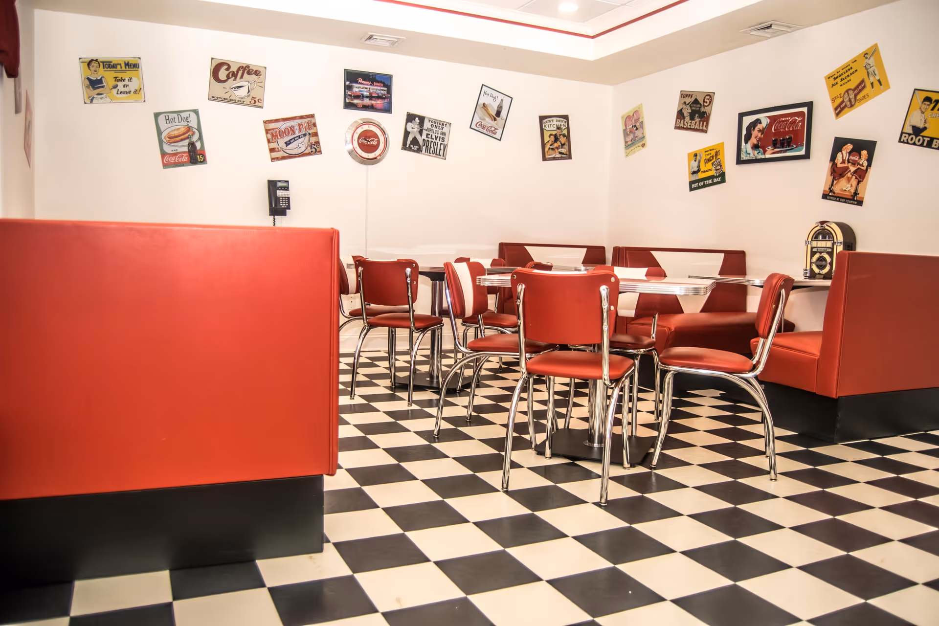 A retro-style dining area with red vinyl booths and chairs, black and white checkered floor tiles, and vintage advertising signs on the white walls, including Coca-Cola and Elvis Presley themed posters.