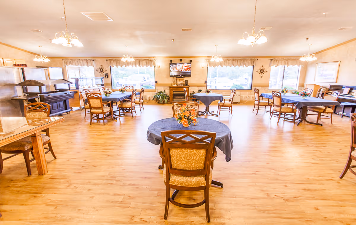 A spacious dining room with multiple round tables covered with blue tablecloths, each adorned with a floral centerpiece. Wooden chairs with cushioned seats surround the tables. The room has large windows letting in natural light, wooden flooring, and several ceiling light fixtures. A television is mounted on the far wall above a small fireplace, and there is a piano on the right side of the room.