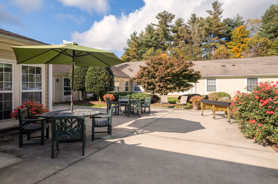 Outdoor patio area at Carolina Reserve of Hendersonville with green patio tables and chairs, a large green umbrella, potted plants, and trees surrounding the concrete seating area under a partly cloudy sky.