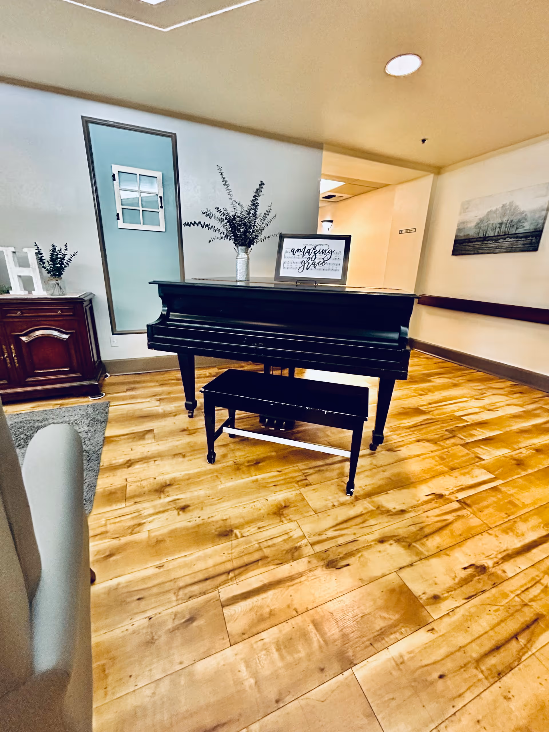 Interior view of a room with a black grand piano and matching bench on a wooden floor. On top of the piano is a vase with greenery and a framed sign that reads 'amazing grace'. To the left, there is a wooden cabinet with decorative items and a small plant. The walls are light-colored with a piece of artwork hanging on the right side.