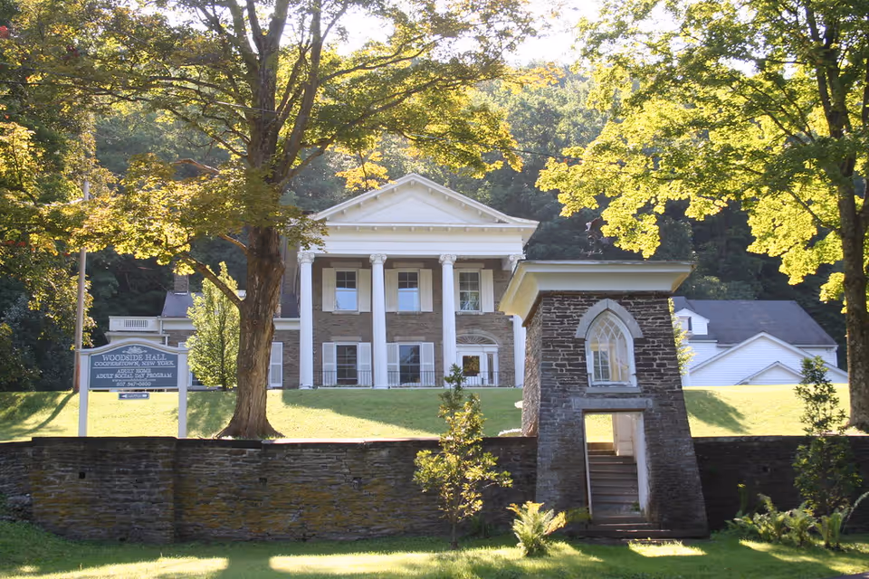 A large historic building with white columns and a triangular pediment, surrounded by trees and greenery. In front of the building is a stone wall with a small arched stone structure containing a staircase. A sign near the building reads 'Woodside Hall, Gooderstown, New York, Adult Home, Adult Social Day Program'.