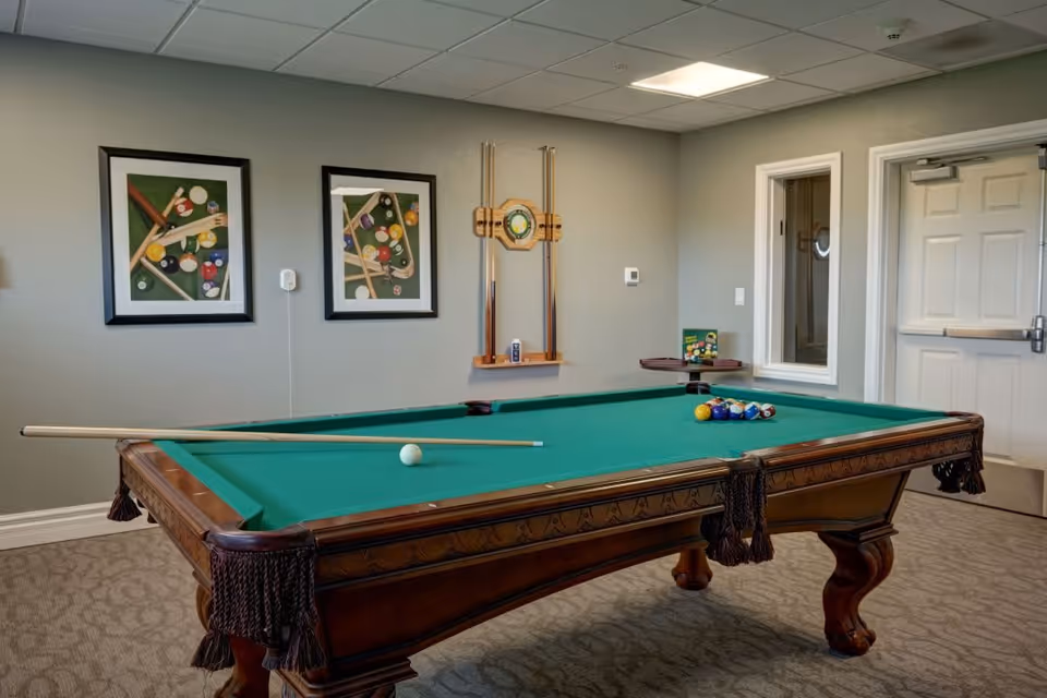 A billiards room with a green pool table featuring a cue stick and billiard balls arranged for a game. The room has beige walls with two framed pictures of pool balls and cues, a wooden rack holding pool cues, and a closed white door in the background.