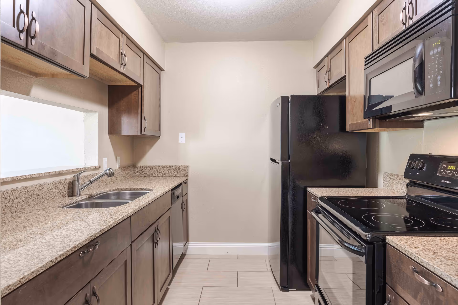 A modern kitchen with granite countertops, dark wooden cabinets, a double sink, a black refrigerator, a black microwave, and a black electric stove. The kitchen has a neutral color scheme with beige walls and tiled flooring.