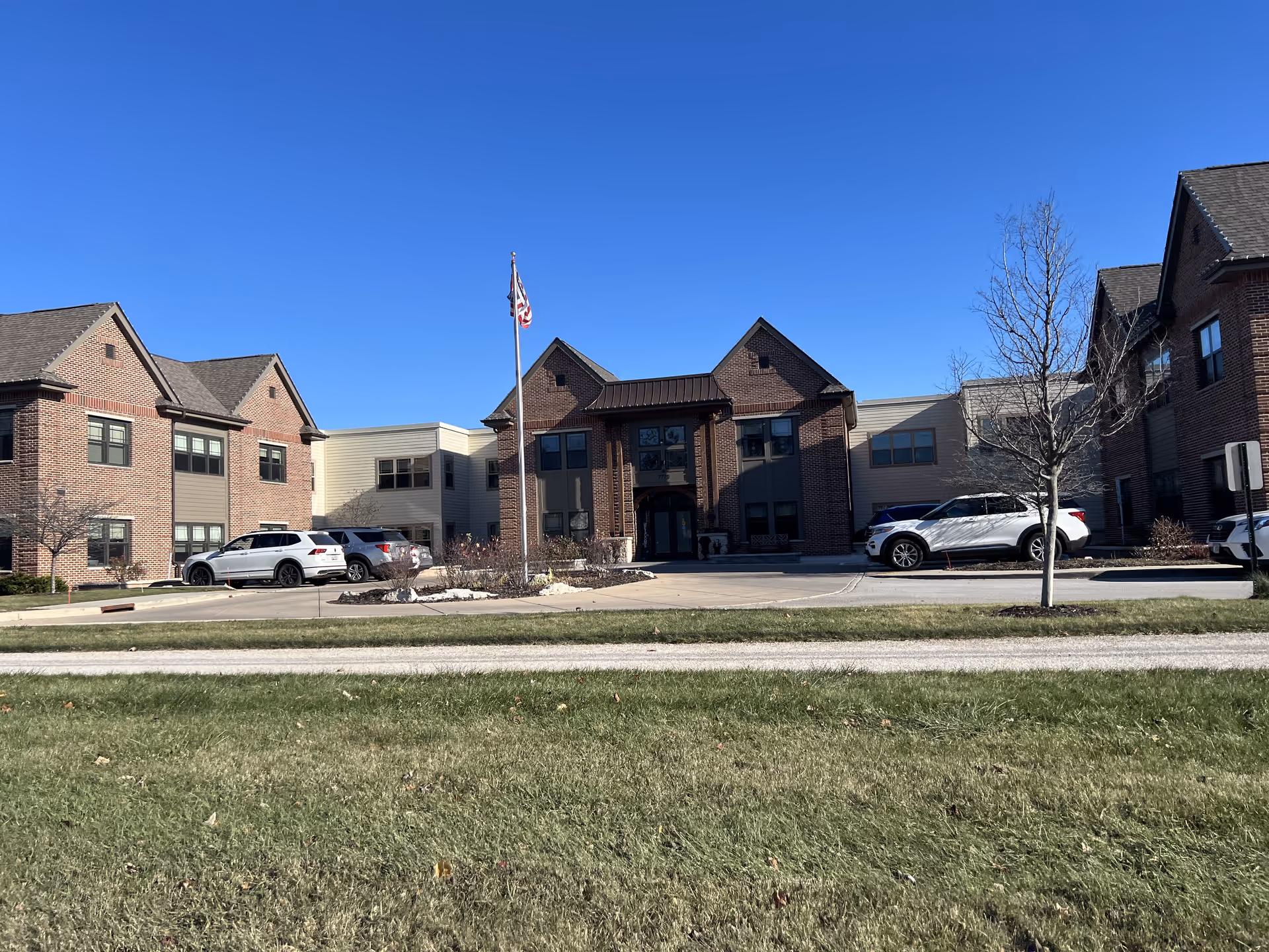 Exterior view of a brick and siding assisted living facility building under a clear blue sky. Several cars are parked in front of the building, and an American flag is flying on a flagpole near the entrance. There is a grassy area in the foreground.