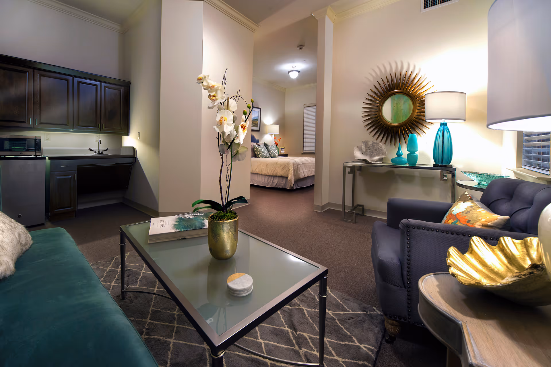 Interior view of a senior living facility suite showing a living area with a green sofa, glass coffee table with a potted orchid, and a blue armchair with a decorative pillow. In the background, there is a small kitchenette with dark cabinets and a bedroom with a bed, nightstand, and lamp. The room is warmly lit with decorative elements including a sunburst mirror and table lamps.
