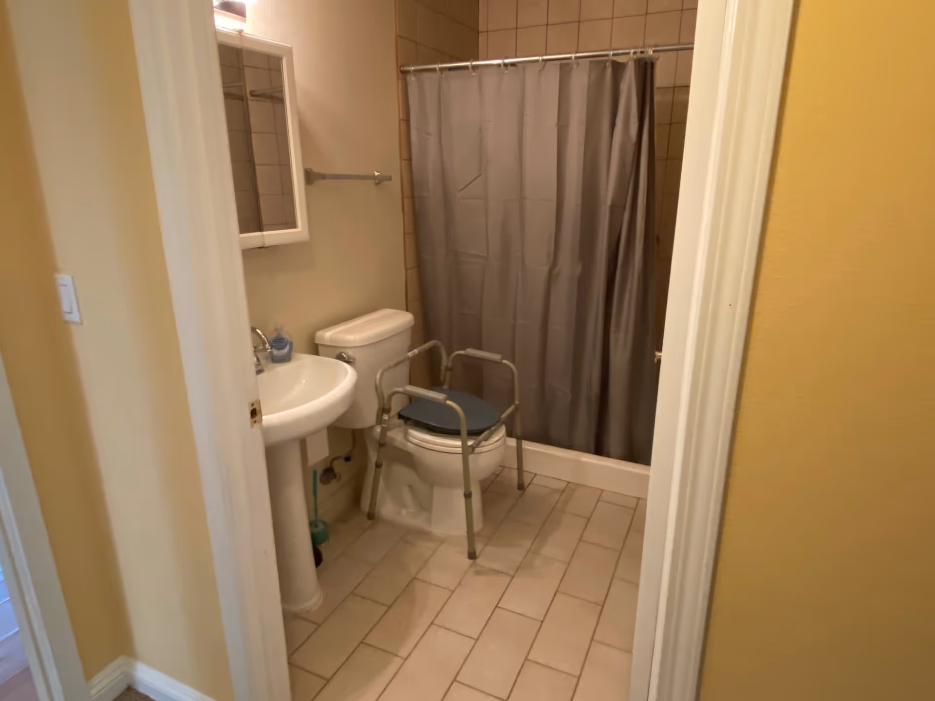 View into a bathroom with beige tiled floor and walls painted yellow and beige. The bathroom contains a white pedestal sink, a toilet with a gray padded toilet safety frame, a gray shower curtain, and a wall-mounted mirror cabinet above the sink.