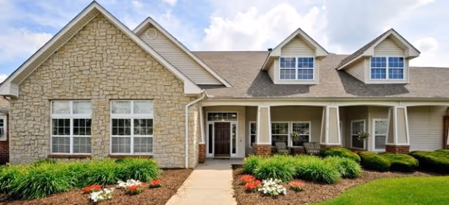 Front exterior of a single-story stone and siding senior living building with a covered porch, dormer windows, walkway, and landscaped shrub beds.