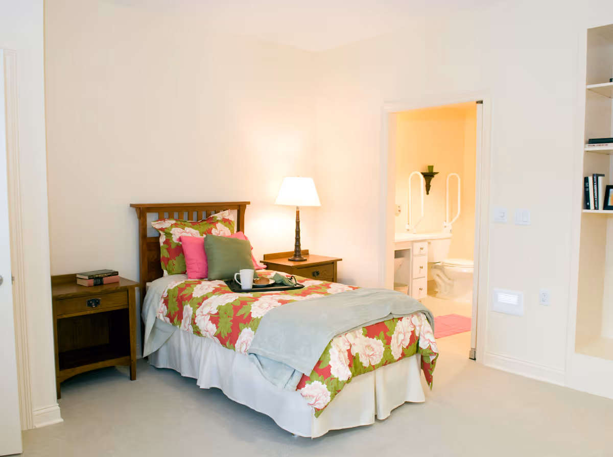 A cozy bedroom with a single bed featuring floral bedding and multiple pillows. A wooden nightstand with a lamp and books is beside the bed. An open door reveals a bathroom with a toilet and grab bars. The room has light-colored walls and carpeted floor.