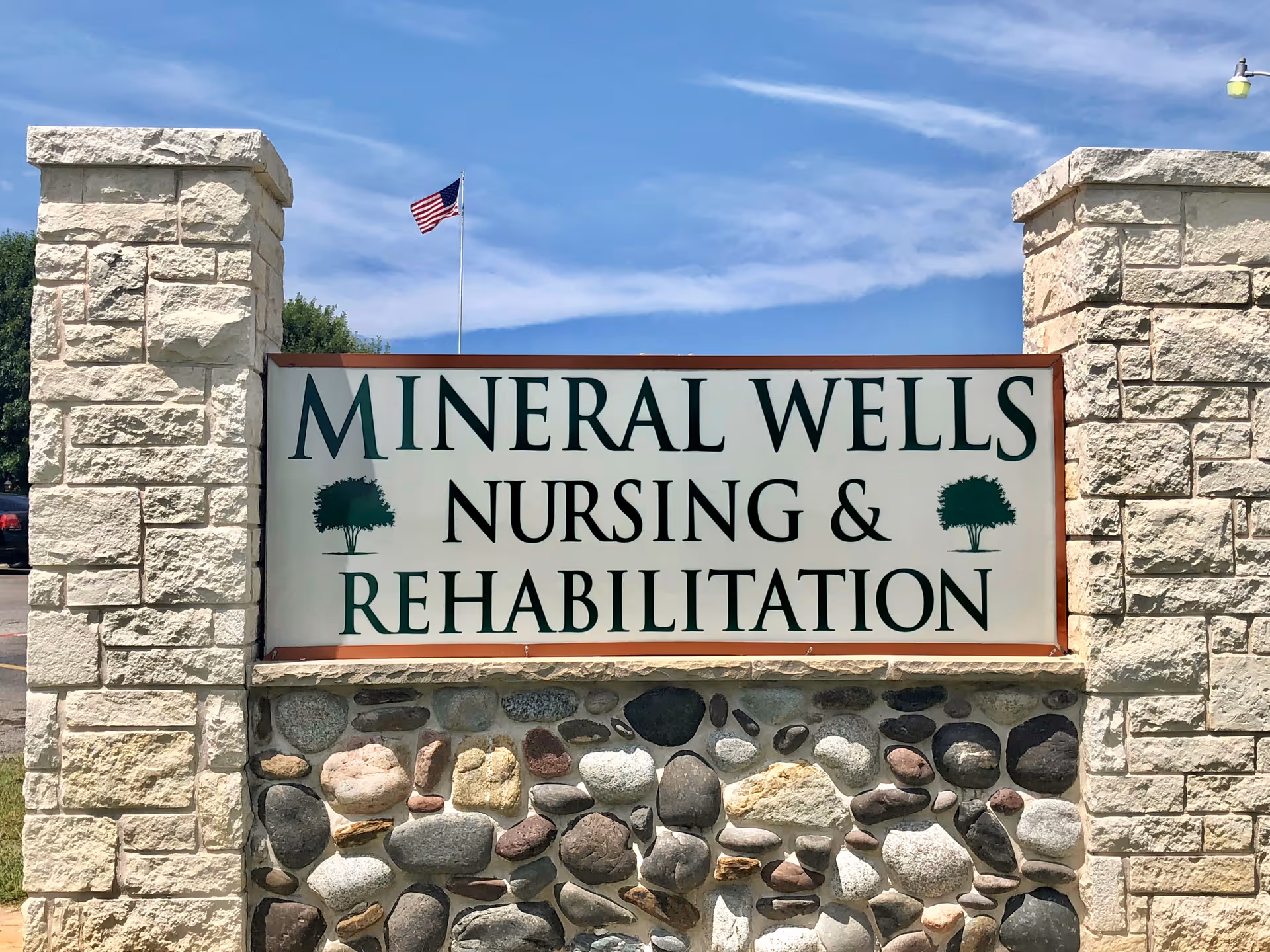 Stone-framed sign that reads "Mineral Wells Nursing & Rehabilitation" with an American flag and blue sky in the background.