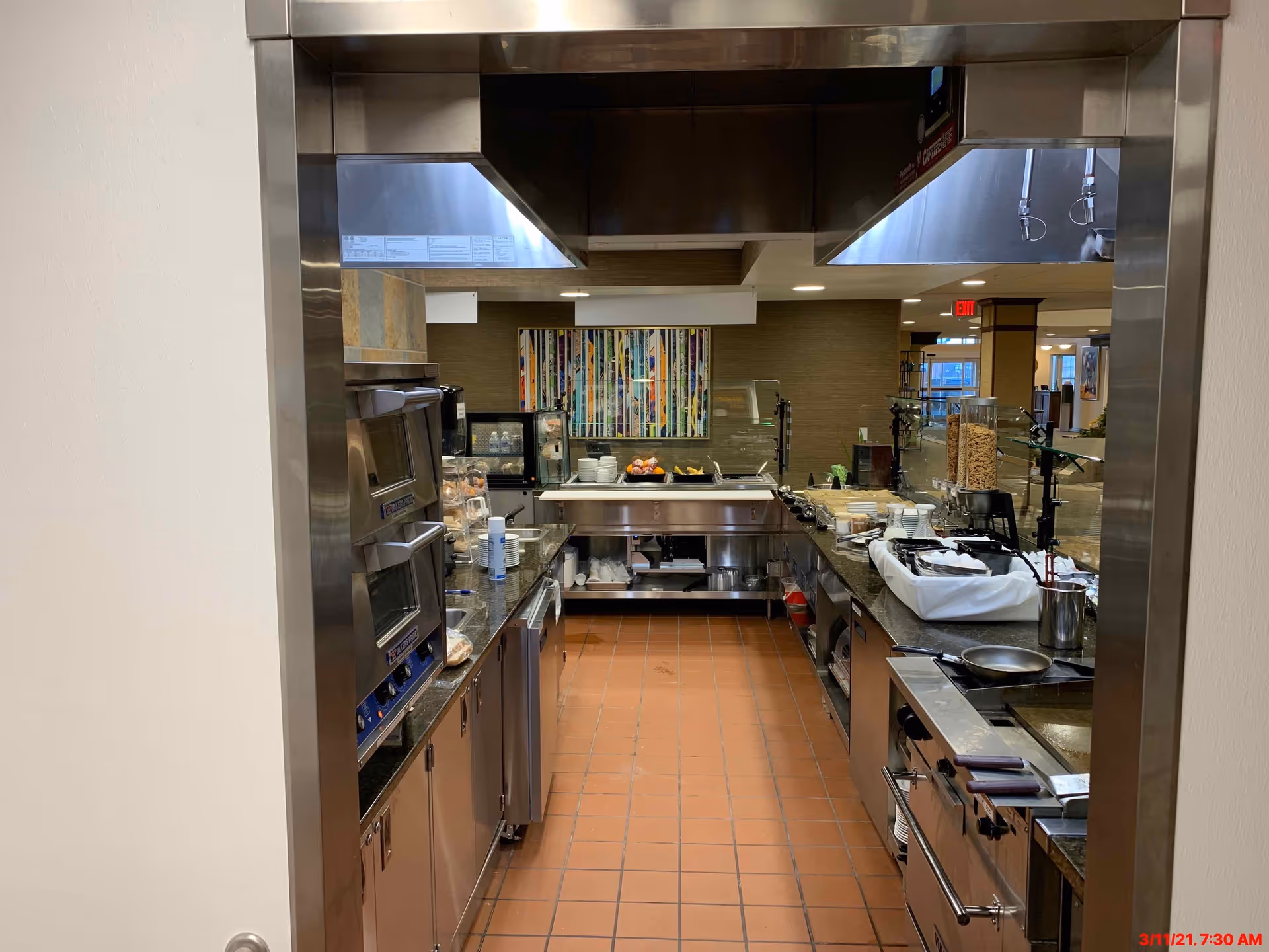 View into a commercial kitchen area with stainless steel appliances, ovens, and countertops. The kitchen has a tiled floor and is equipped with various cooking utensils and food items. In the background, there is a colorful abstract painting on the wall and a glimpse of a dining or common area beyond the kitchen.