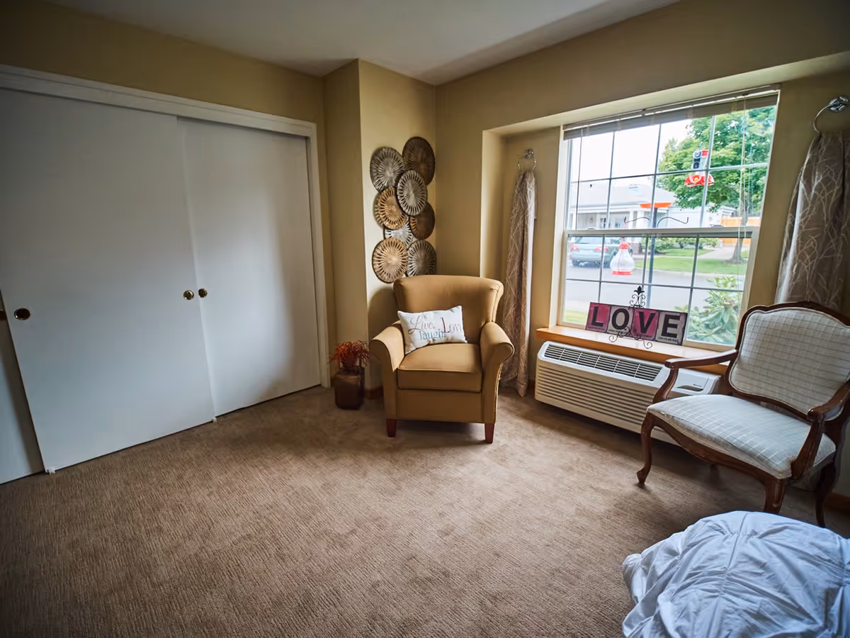 A cozy room with beige walls and carpet, featuring a tan armchair with a decorative pillow, a wooden framed upholstered chair, a large window with curtains, and a white air conditioning unit below the window. Decorative metal wall art is hung on the corner wall, and a decorative sign spelling 'LOVE' is displayed on the window sill.