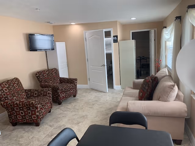 A cozy living room area in an assisted living facility with two patterned armchairs, a beige sofa with decorative pillows, a wall-mounted flat-screen TV, and a small black table with chairs in the foreground. The room has beige walls, tiled flooring, and two open doors leading to other rooms.