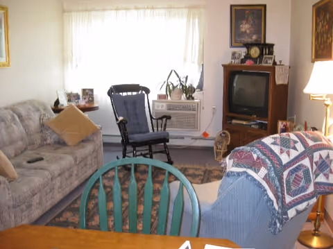 A cozy living room with a patterned sofa and pillows on the left, a wooden rocking chair with a cushion in front of a window with sheer curtains, a TV on a wooden stand with framed pictures and a clock on top, a blue armchair with a quilt draped over the back, a green wooden chair in the foreground, and a floor lamp on the right side.