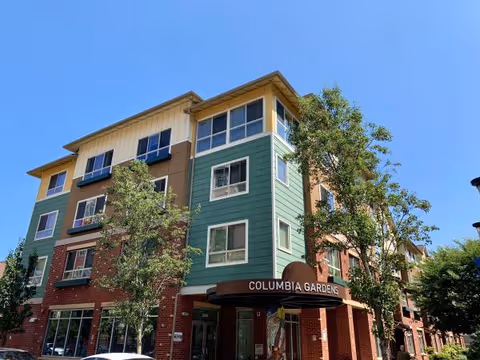 Exterior front of a multi-story senior living building with a 'Columbia Gardens' awning, trees, and a clear blue sky.