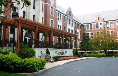 Front exterior of SEM Villa showing a brick multi-story building with a covered pergola entrance, landscaping, benches, and a driveway.