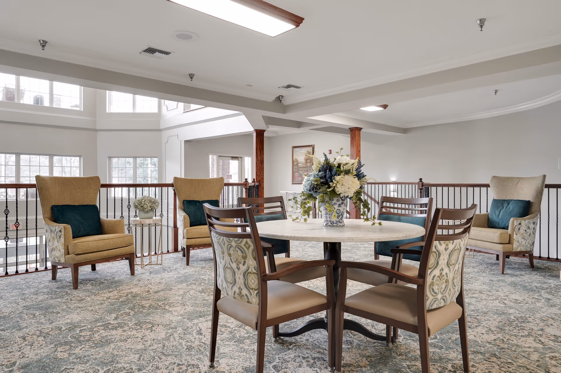 A bright and spacious common area with a round table and four chairs in the center, decorated with a floral arrangement. Surrounding the table are three upholstered armchairs with dark green cushions. The area features large windows, a patterned carpet, and wooden railings overlooking a lower level.