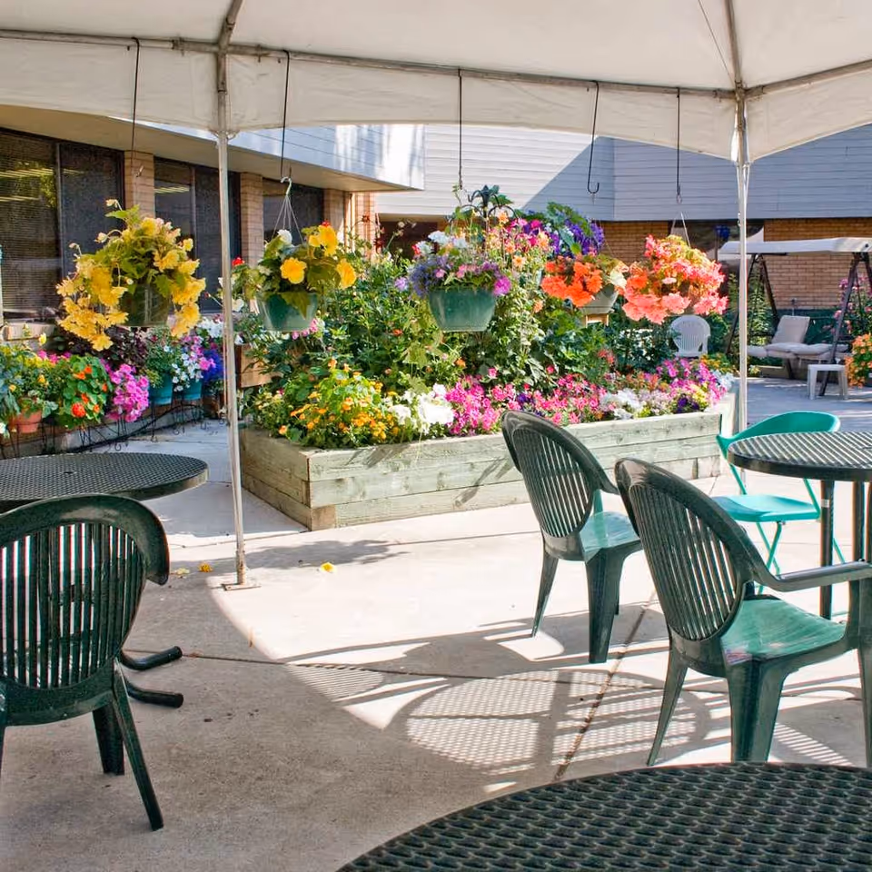 Outdoor patio area with green plastic chairs and round tables under a white canopy. In the background, there is a raised garden bed filled with colorful flowers and several hanging flower pots. The setting is bright and sunny with a building visible behind the garden.