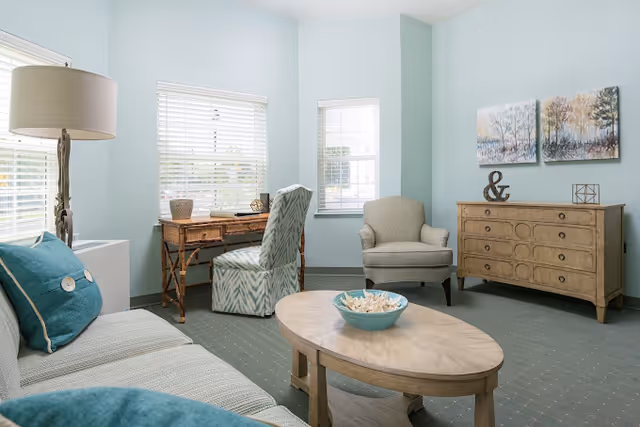 A bright and cozy living room with light blue walls and carpet. The room features a beige sofa with teal pillows, a wooden oval coffee table with a decorative bowl, a wooden desk with a patterned chair, a beige armchair, and a wooden dresser with decorative items and two landscape paintings above it. Large windows with white blinds allow natural light to fill the space.