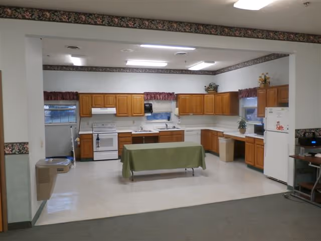 A large kitchen area with wooden cabinets, a white stove, refrigerator, and sink. A table covered with a green cloth is placed in the center. The room has white walls with floral wallpaper borders near the ceiling and windows with maroon valances. There is a water fountain on the left and a small desk with electronic equipment on the right.