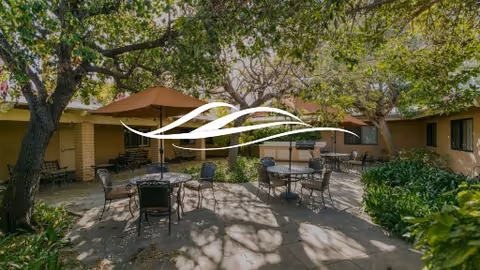 Outdoor patio area at Mainplace Senior Living with several metal tables and chairs arranged under large brown umbrellas, surrounded by trees and greenery, with a building visible in the background.