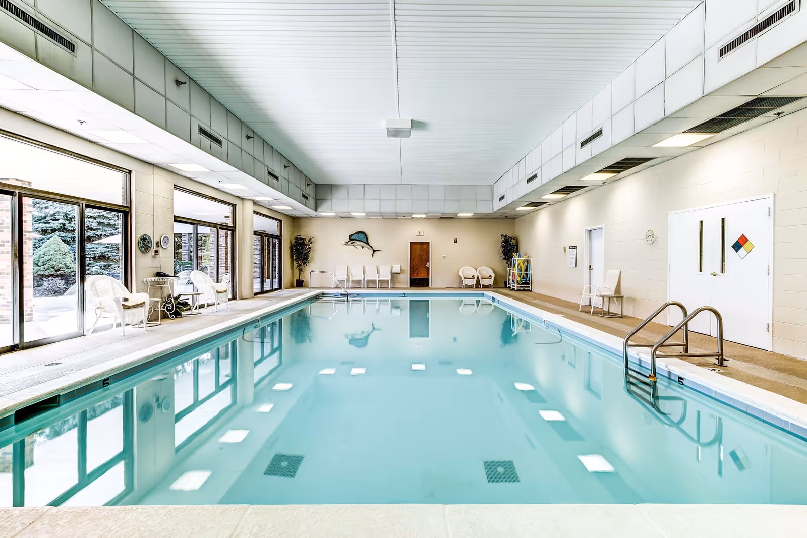 Indoor swimming pool with clear blue water surrounded by white walls and large windows on one side. White chairs and plants are placed around the pool area, and there is a door at the far end with a fish decoration mounted on the wall above it.