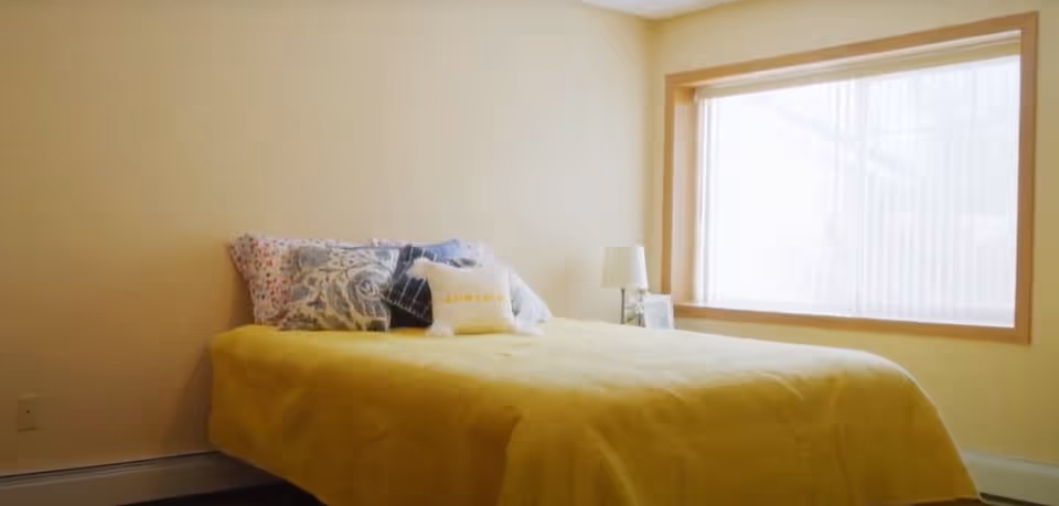 A bedroom with a bed covered in a yellow blanket and several decorative pillows. There is a small bedside table with a lamp and a framed photo next to a large window with vertical blinds.