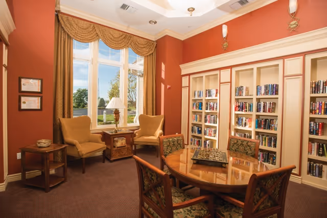A cozy reading and sitting area with two upholstered armchairs near a large window with gold curtains. A wooden side table with a lamp is placed between the chairs. Opposite the chairs is a round wooden table with four chairs, featuring a chessboard on top. Built-in bookshelves filled with books line the wall behind the table. The walls are painted a warm reddish color, and the room is carpeted.