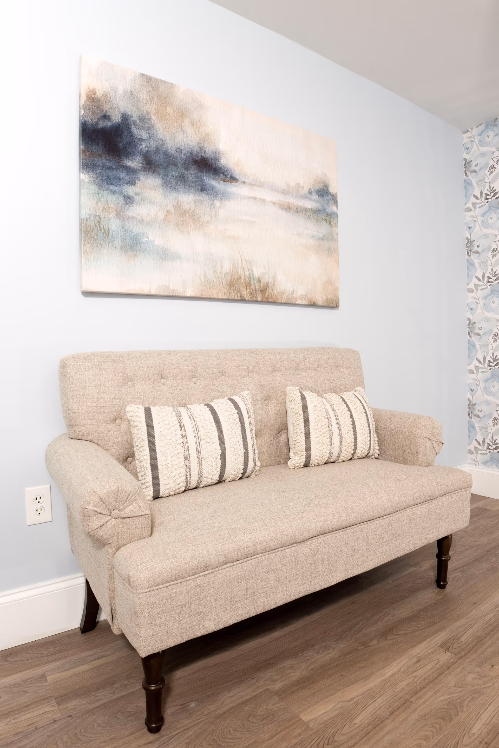 A beige upholstered loveseat with two textured striped pillows placed against a light blue wall. Above the loveseat hangs a large abstract painting with soft blue, beige, and white tones. The floor is wood, and part of a floral patterned wallpaper is visible on the right side.