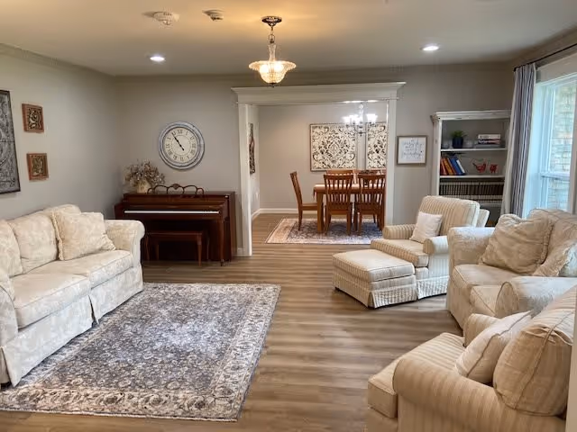 A cozy living room with beige upholstered sofas and armchairs arranged around a patterned area rug on wooden flooring. A piano with a bench is against the far wall beneath a large round clock. The room opens into a dining area with a wooden dining table and chairs, illuminated by a chandelier. A bookshelf with books and decorative items is near a window with curtains.