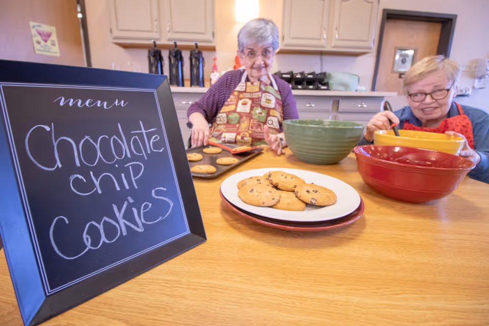 Two elderly women wearing aprons are baking chocolate chip cookies in a kitchen. One woman is placing a tray of cookies on the counter, while the other is stirring ingredients in a yellow bowl. A plate of freshly baked cookies is on the table, and a chalkboard sign reads 'menu Chocolate chip Cookies'.