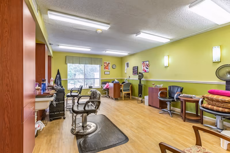 Bright salon room with hairdressing chairs, workstations, and salon equipment against green walls and a large window.