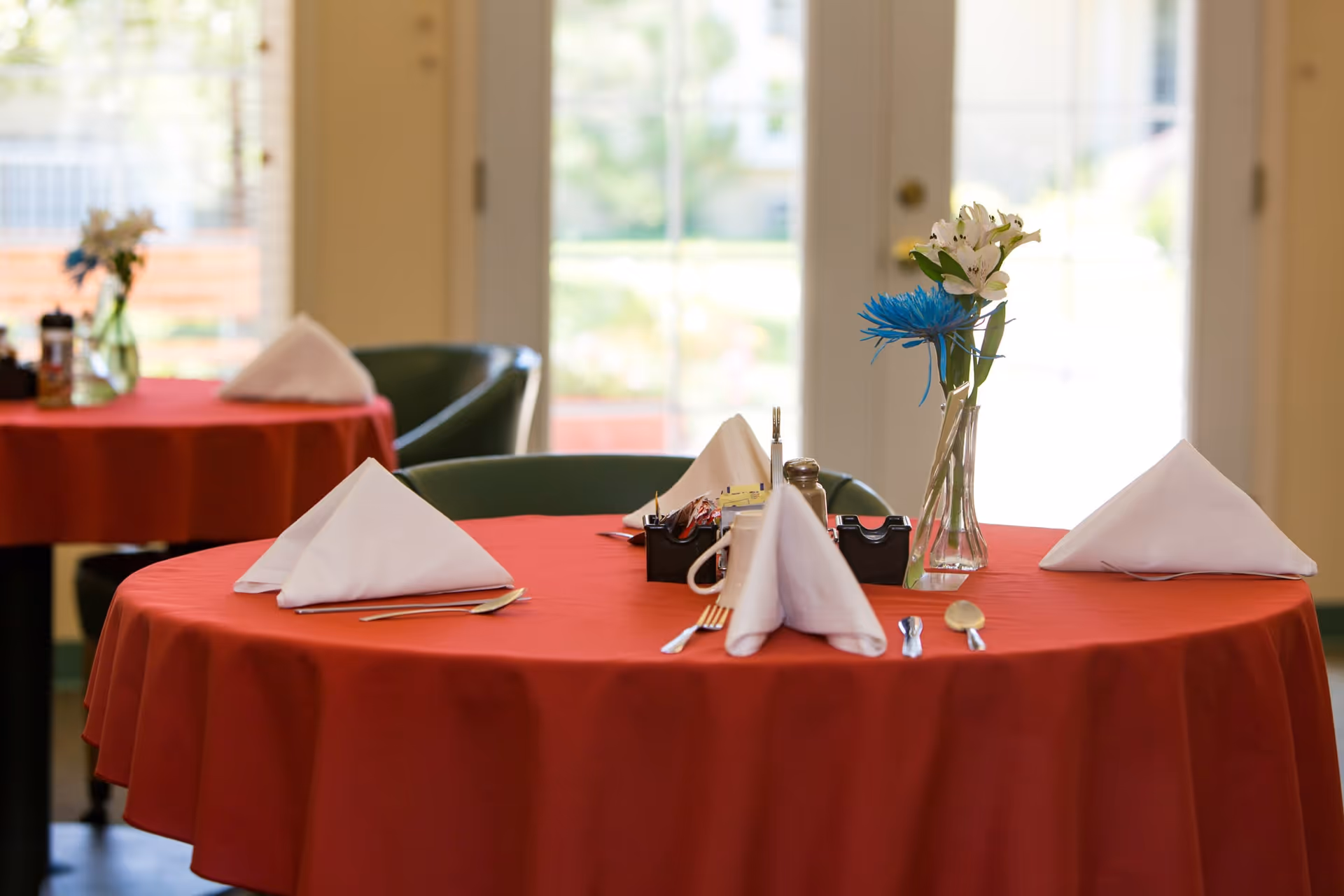 A dining table set with a red tablecloth, white folded napkins, silverware, a small condiment holder, and a vase with white and blue flowers. In the background, another similarly set table is visible near a window with natural light coming through.