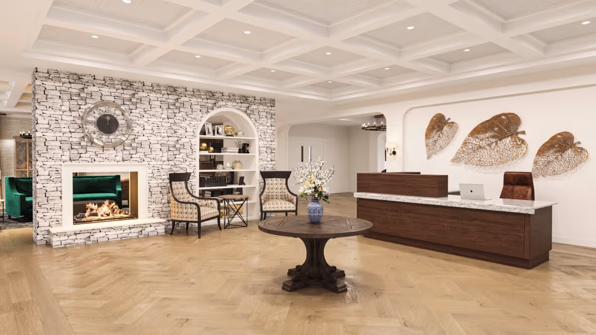 A spacious and elegant reception area with a wooden reception desk on the right, decorated with three large metallic leaf wall art pieces above it. In the center, there is a round wooden table with a blue vase holding white flowers. To the left, there is a stone accent wall with a modern fireplace and two patterned armchairs with a small table between them. The floor is light wood with a herringbone pattern, and the ceiling features a coffered design with recessed lighting.
