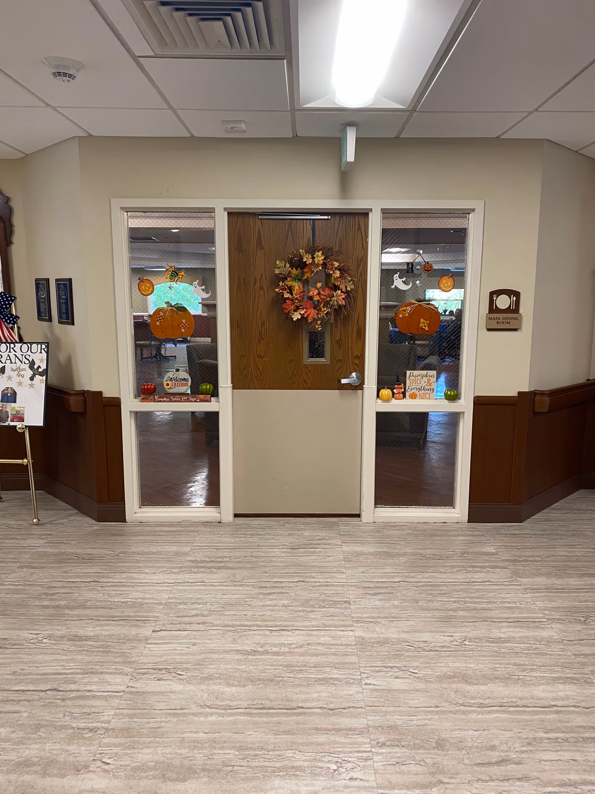 A hallway leading to a wooden door decorated with an autumn-themed wreath. The door is flanked by two glass panels with fall decorations including pumpkins and ghost cutouts. A sign on the right side of the door reads 'Main Dining Room'. The floor is light-colored tile and the walls are beige with wooden paneling on the lower half.