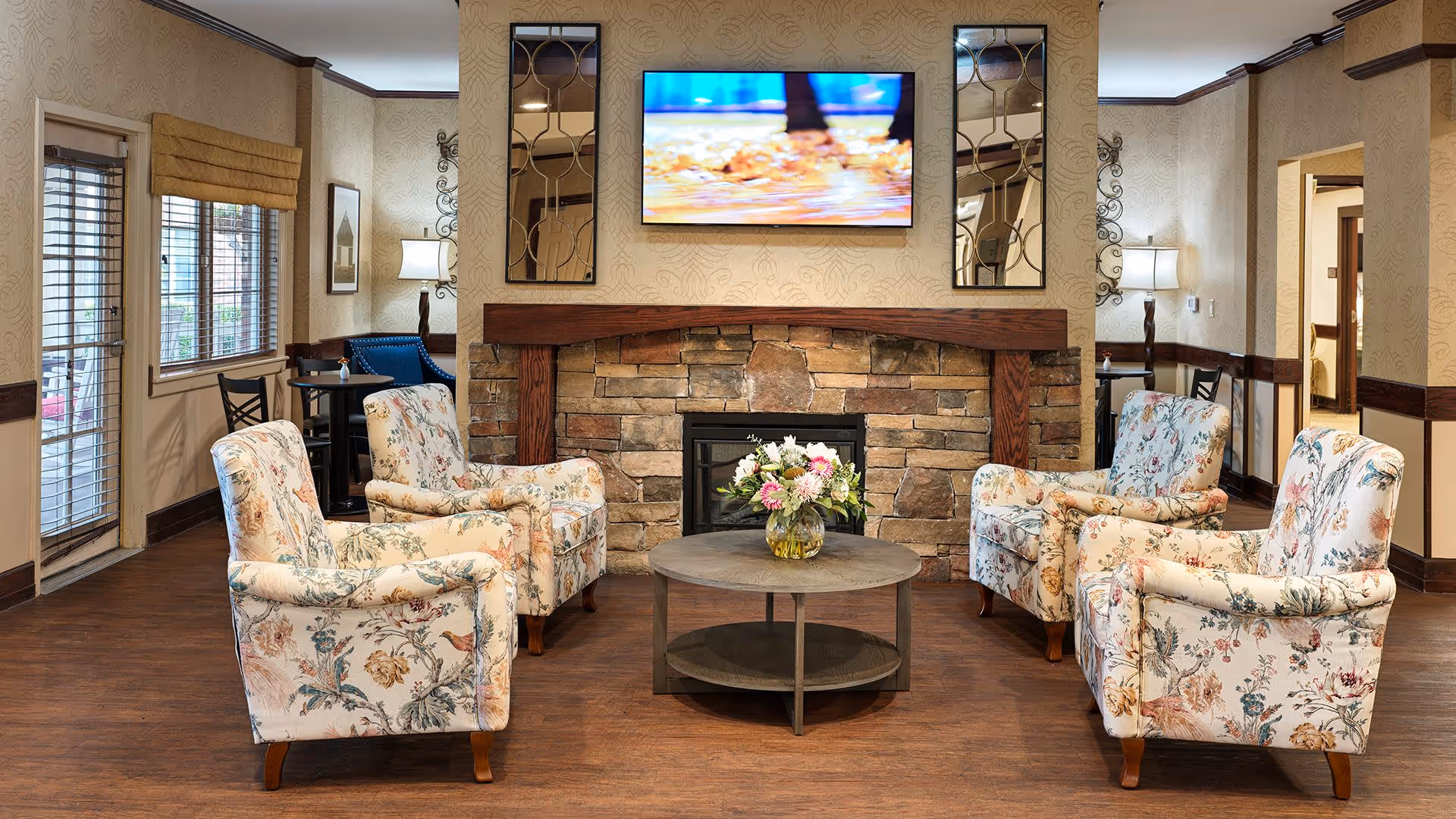 A cozy living room area with four floral upholstered armchairs arranged around a round wooden coffee table with a vase of flowers. Behind the chairs is a stone fireplace with a wooden mantel, above which hangs a flat-screen TV. The room has wooden floors, beige patterned wallpaper, and windows with blinds and a yellow valance. There are also two decorative mirrors flanking the TV and a small seating area with a table and chairs near the window.