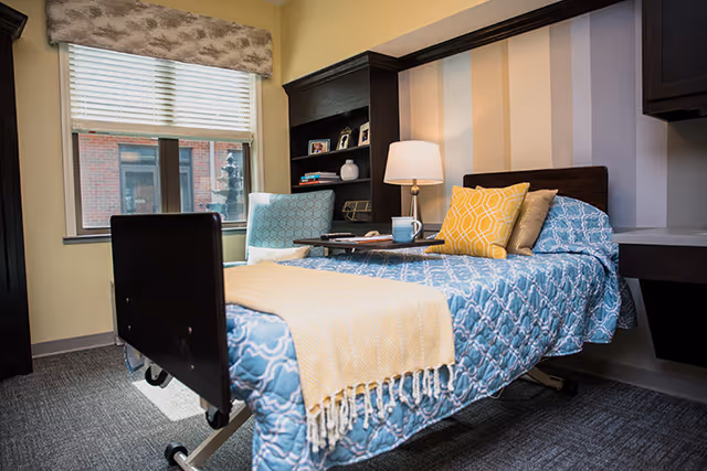 A cozy bedroom in a senior living facility featuring a single bed with a blue patterned bedspread, yellow and beige pillows, and a beige throw blanket. Next to the bed is a wooden nightstand with a lamp and a tray holding a cup. A comfortable armchair is positioned near a window with blinds and a valance. A built-in bookshelf with framed photos and decorative items is visible behind the chair.