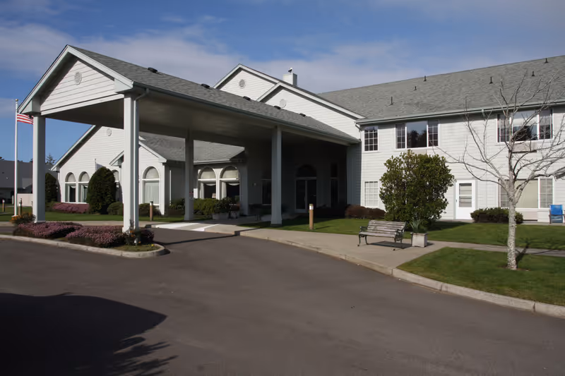 Front entrance of a light-colored assisted living building with a covered porte-cochere, driveway, benches, and landscaped lawn.