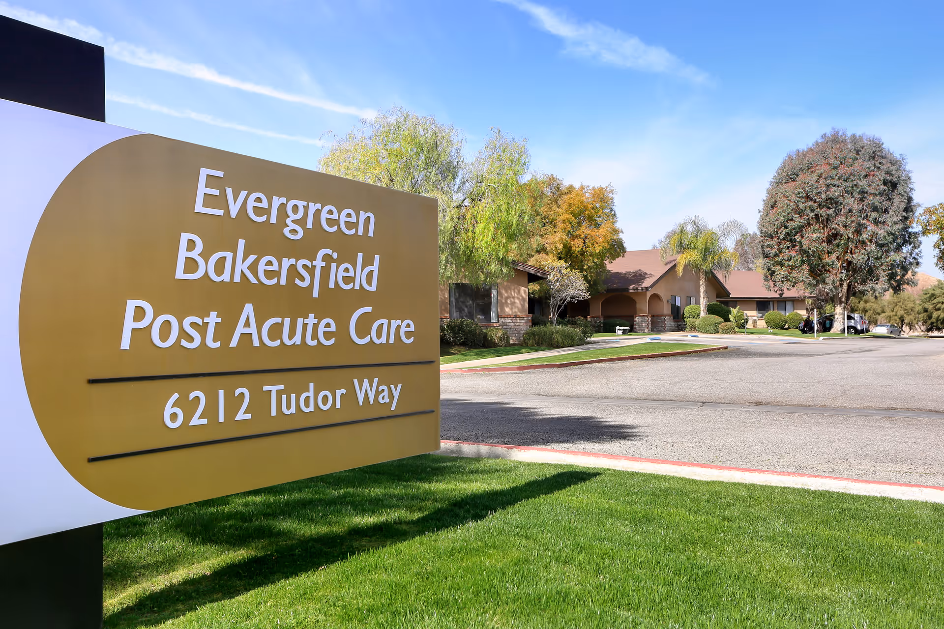 Outdoor view of Evergreen Bakersfield Post Acute Care facility sign with the address 6212 Tudor Way, showing a driveway, green lawn, trees, and a building in the background under a blue sky.