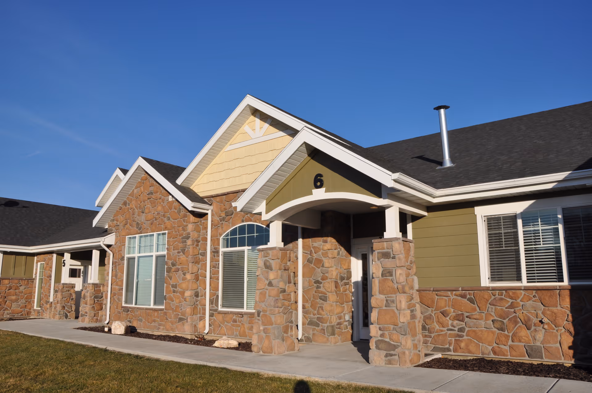 Exterior view of a single-story senior living facility building with stone and green siding, numbered units 5 and 6 visible, under a clear blue sky.