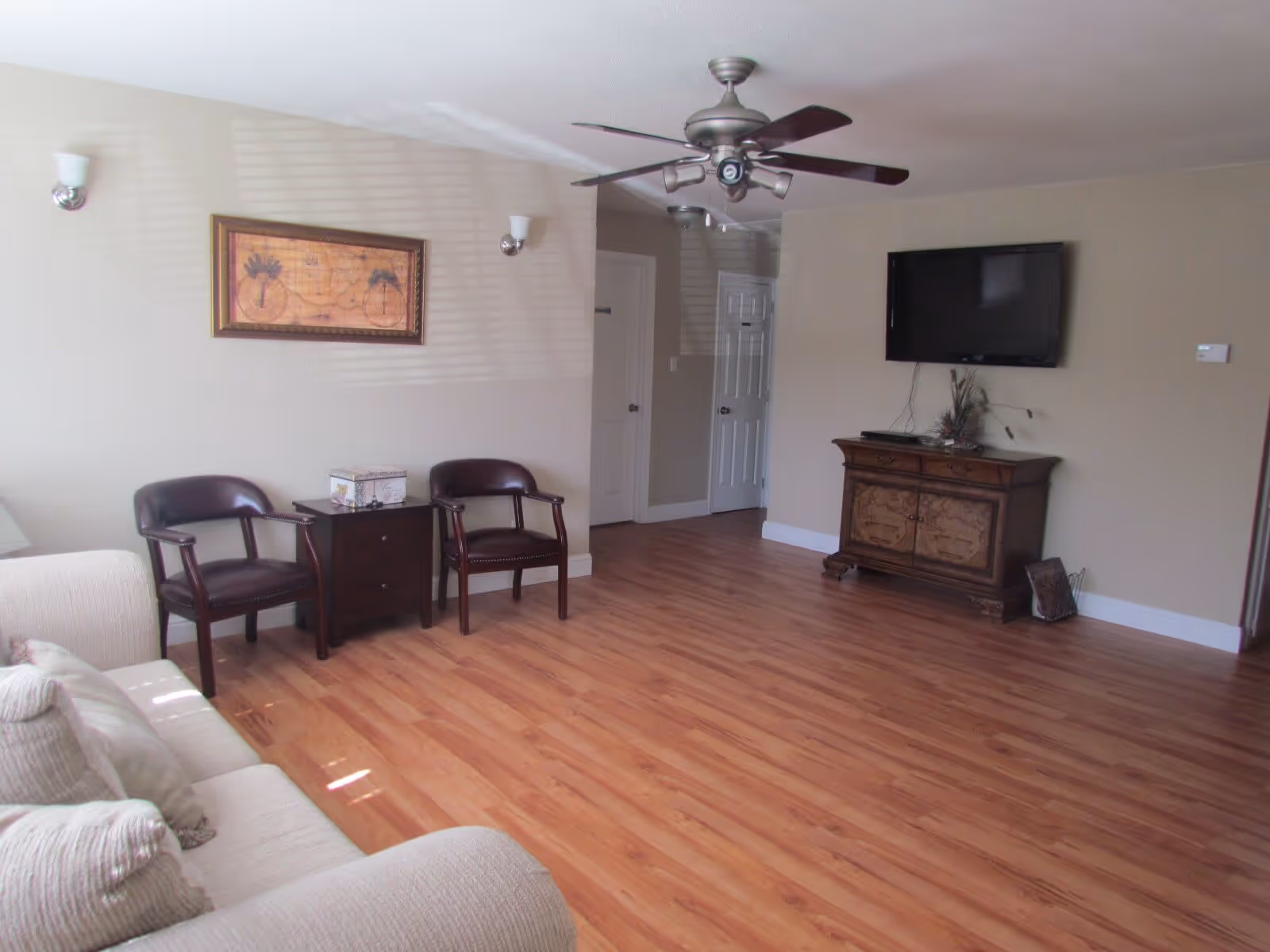 A living room with wooden flooring, a beige couch with cushions on the left, two dark wooden chairs with a small dark wooden side table between them against the wall, a ceiling fan with lights, a wall-mounted flat-screen TV above a wooden cabinet, and a framed picture hanging on the wall.