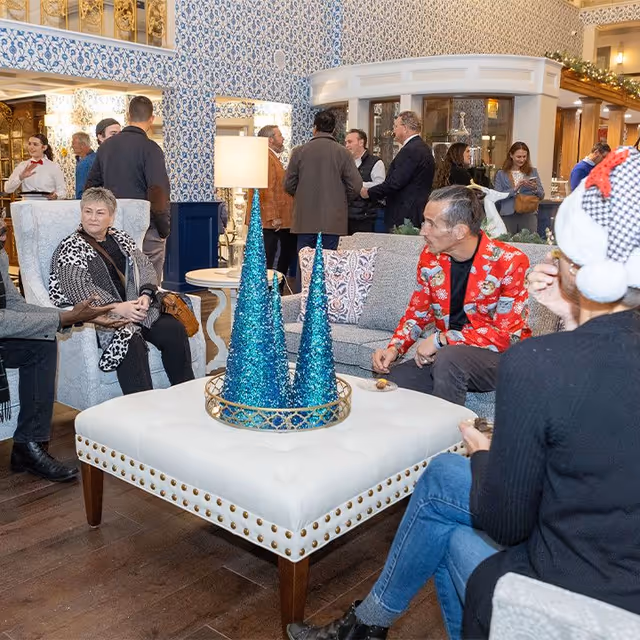 A festive indoor lounge area with several people socializing. The room features patterned wallpaper, a large white ottoman with two blue glittery cone-shaped decorations, and comfortable seating including armchairs and a sofa. Some people are seated while others stand and chat in the background. One person is wearing a red holiday-themed jacket and another is wearing a Santa hat.