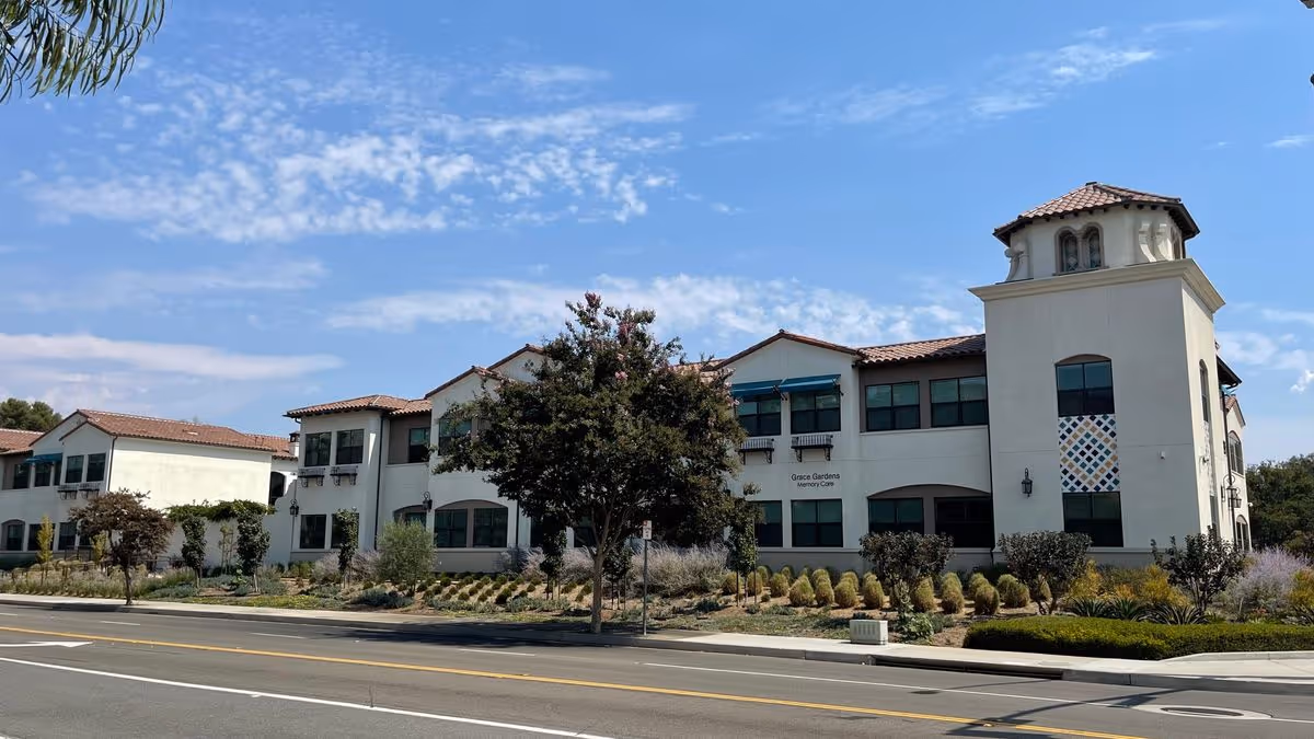 Two-story Mediterranean-style senior living building with a tower, landscaped front yard, and a street in the foreground.