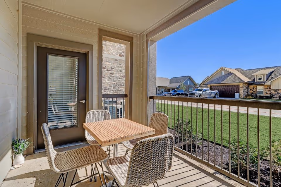 Covered balcony with a small wooden table and four wicker chairs overlooking a residential street and front yards.