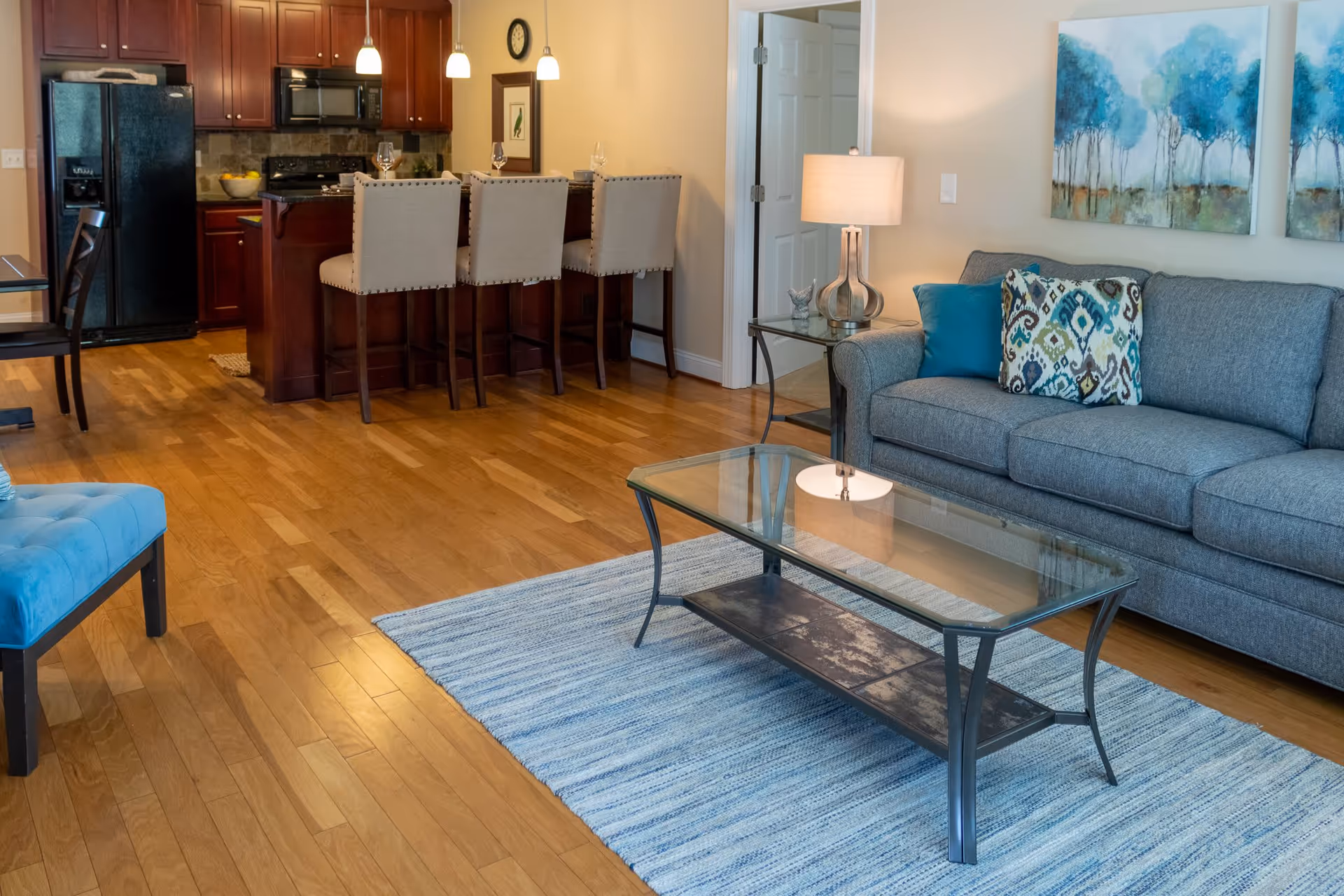 Interior view of a senior living facility showing a living room and kitchen area. The living room has a gray sofa with patterned and solid blue pillows, a glass coffee table on a blue striped rug, and a side table with a lamp. The kitchen features dark wood cabinets, a black refrigerator, a stove, and a kitchen island with four beige upholstered bar stools. The floor is wooden, and there are two paintings of blue trees on the wall above the sofa.
