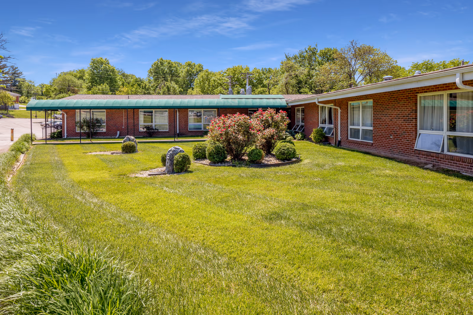 Exterior view of a single-story brick building with large windows, a green awning over the entrance, and a well-maintained lawn with bushes and small trees under a clear blue sky.