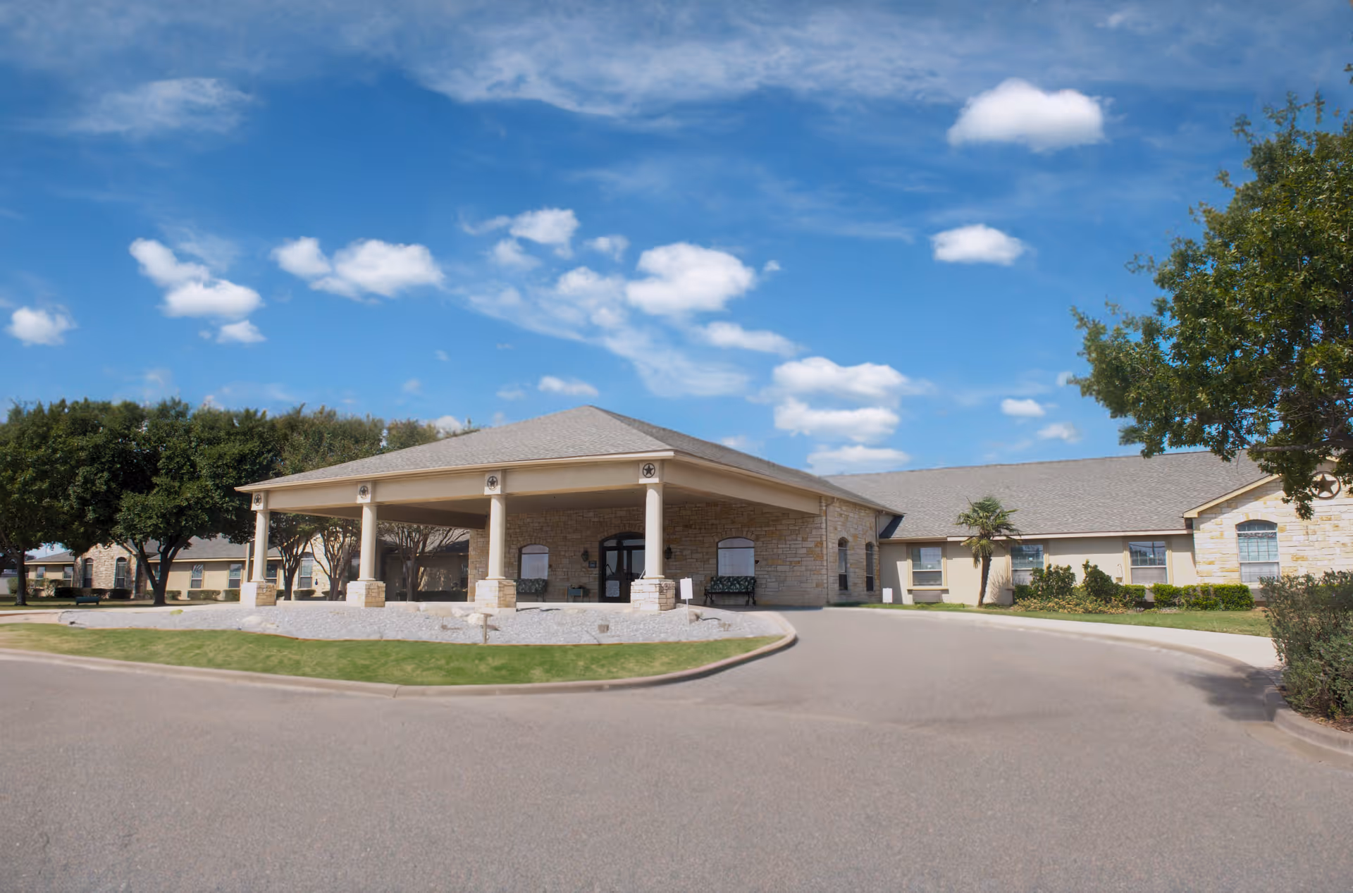 Single-story stone healthcare building front with a covered porte-cochère, circular driveway, trees and blue sky.